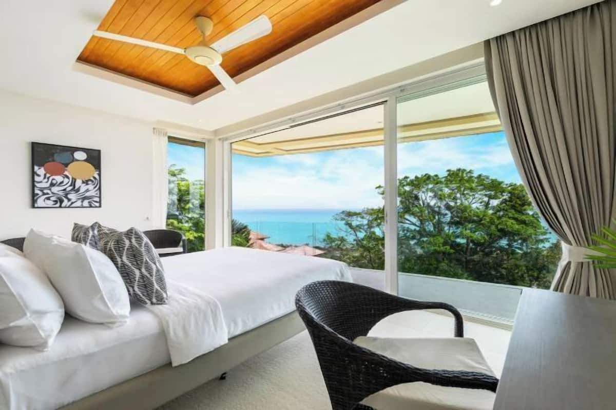 Une chambre à coucher comprenant un lit, une chaise, un ventilateur de plafond et une grande fenêtre donnant sur l’océan.