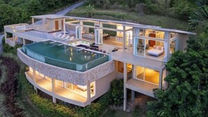 A modern, multi-level house with a large glass-enclosed infinity pool, outdoor seating, and a well-lit interior. - Villa Cabo: Hilltop Retreat with Panoramic Ocean Views (Koh Samui)