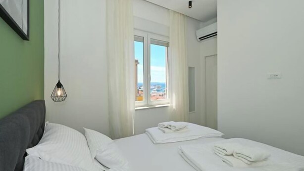 A hotel room with a bed, a window with curtains, a wall-mounted air conditioner, and a wall-mounted light fixture. - Steps from Palace & Riva - Old Town Pine (Split)