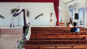 Een kerkinterieur met houten banken, een podium met bloemversiering en een rood gordijn.