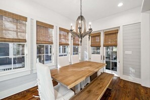 Une salle à manger avec une table en bois et des chaises blanches, de grandes fenêtres équipées de stores et un lustre.