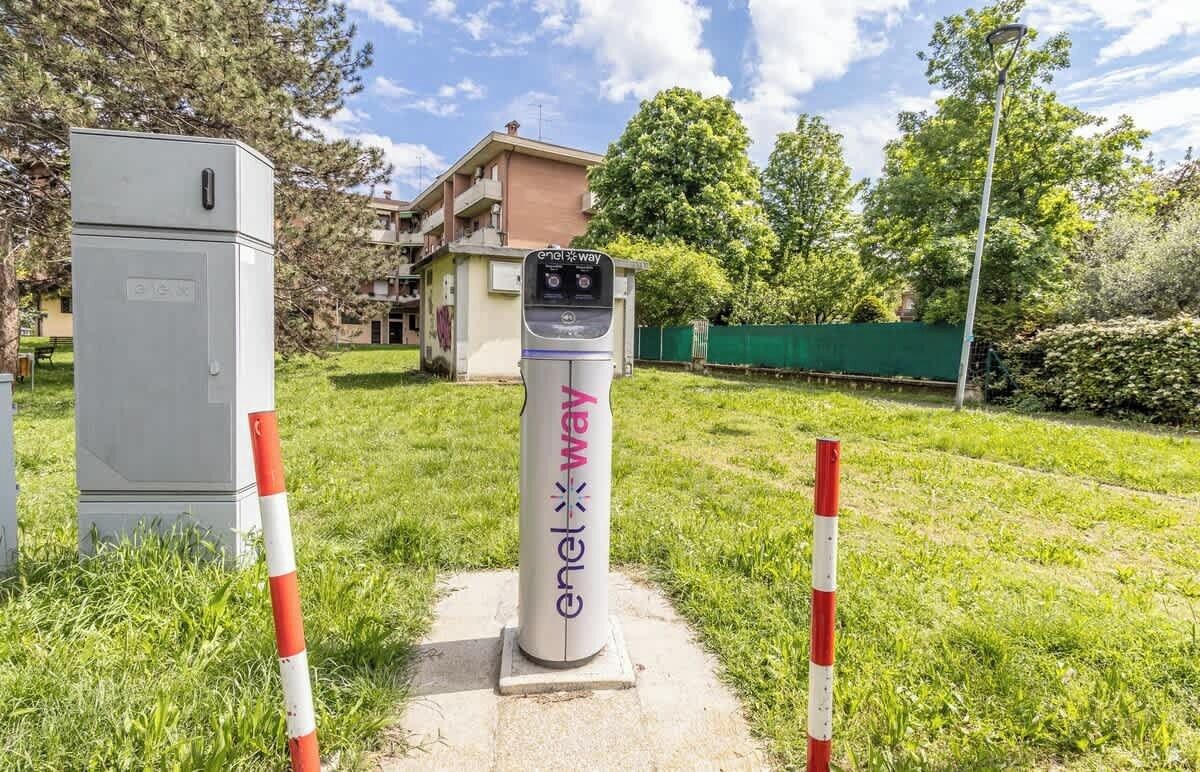 An electric vehicle charging station labeled 'enel way' in a grassy area with a building and trees in the background.