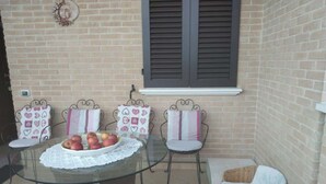 Een terras met een ronde glazen tafel, twee rieten stoelen en een rieten bank.