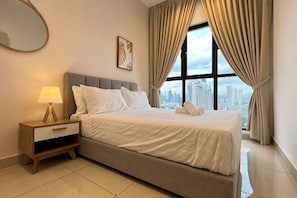 Una habitación de hotel con cama, mesita de noche, lámpara, ventana con cortinas y vista a la ciudad.