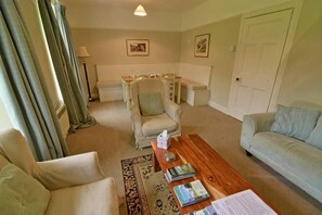 Een woonkamer met een bank, fauteuil, salontafel en een stoel. Er is een lamp, een deur en een raam met gordijnen.