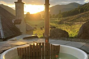 Un jacuzzi con estructura de madera, una chimenea y vista a un paisaje rural con montañas y animales pastando.