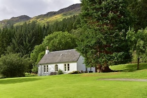 A white building with a sloped roof, surrounded by a green lawn and trees, with mountains in the background. - Gardener's Cottage, Glen Lyon (Aberfeldy)