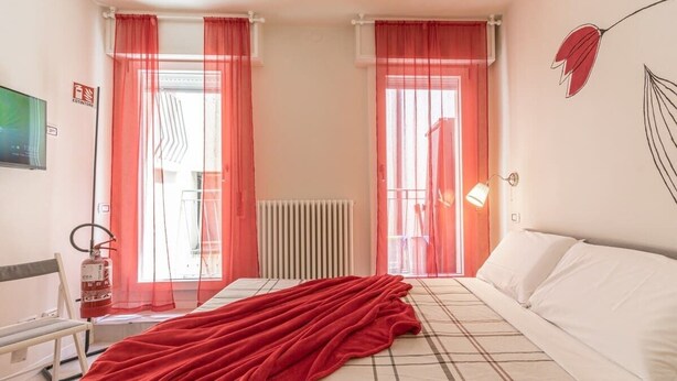 A bedroom with a bed, a red blanket, a radiator, a fire extinguisher, and a window with curtains. - Suite Modena al Centro - Affitti Brevi Italia (Modena)