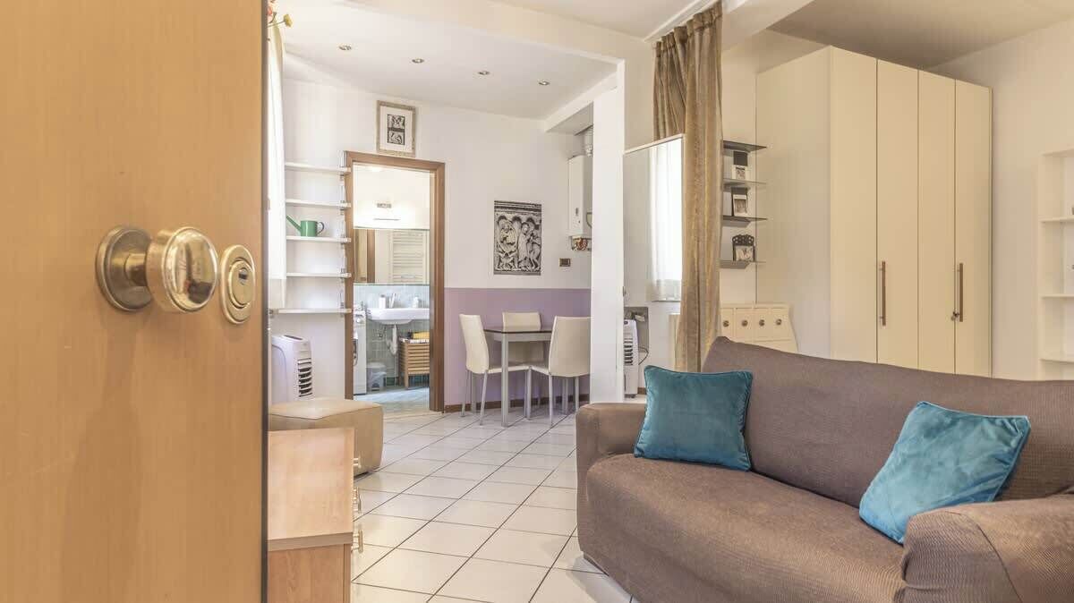A modern living room with a sofa, a dining area with chairs, and a kitchen visible through an open door.