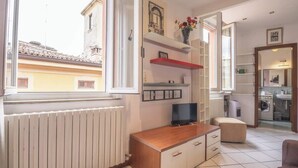 A well-lit room with a wooden cabinet, a television, a red shelf, and a window overlooking buildings. - MonoliVi 35 - Affitti Brevi Italia (Modena)