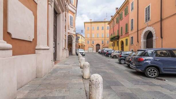 A narrow street lined with parked cars and historic buildings with arched doorways. - MonoliVi 35 - Affitti Brevi Italia (Modena)