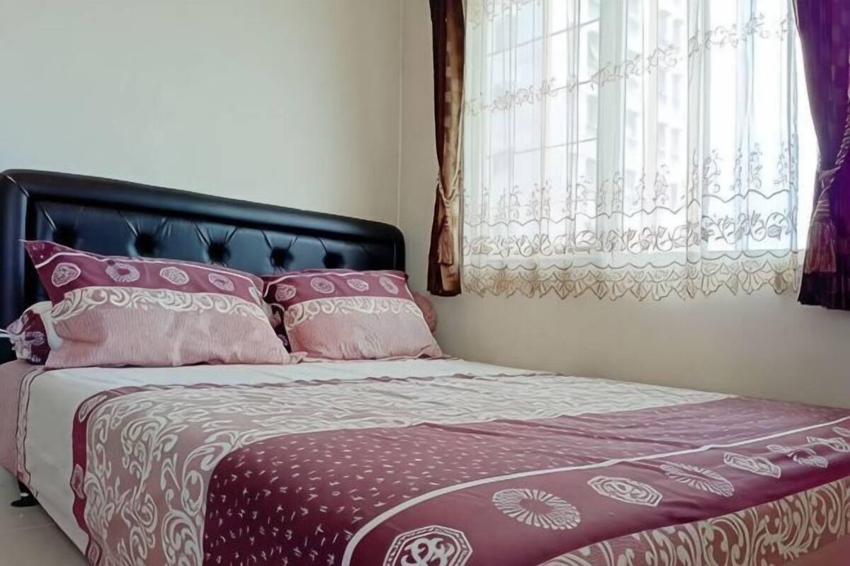 A bed with a patterned bedspread, a dark headboard, and a window with curtains.