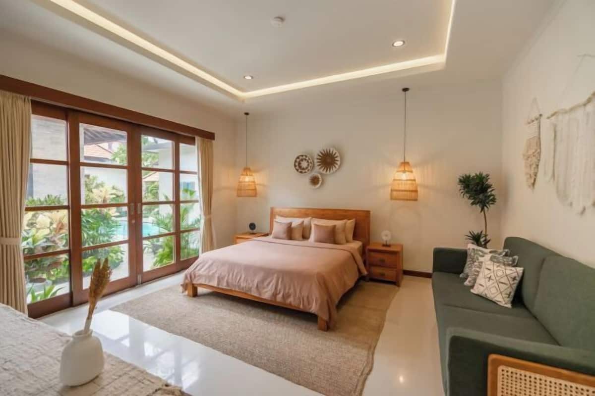 A bedroom with a bed, bedside tables, a green sofa, and a view of greenery through large windows.