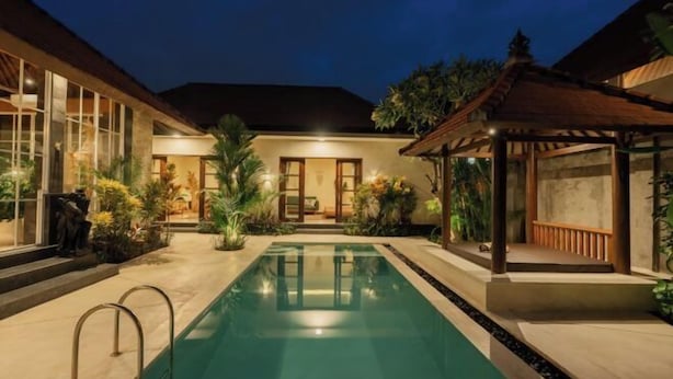 A swimming pool with a wooden deck, surrounded by tropical plants and a traditional gazebo. - Villa Nana by Pertama Stay (Bali)