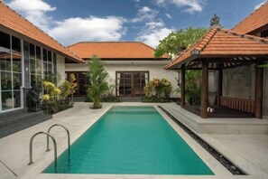 A swimming pool with a tiled deck, surrounded by a building with a tiled roof and large windows. - Villa Nana by Pertama Stay (Bali)