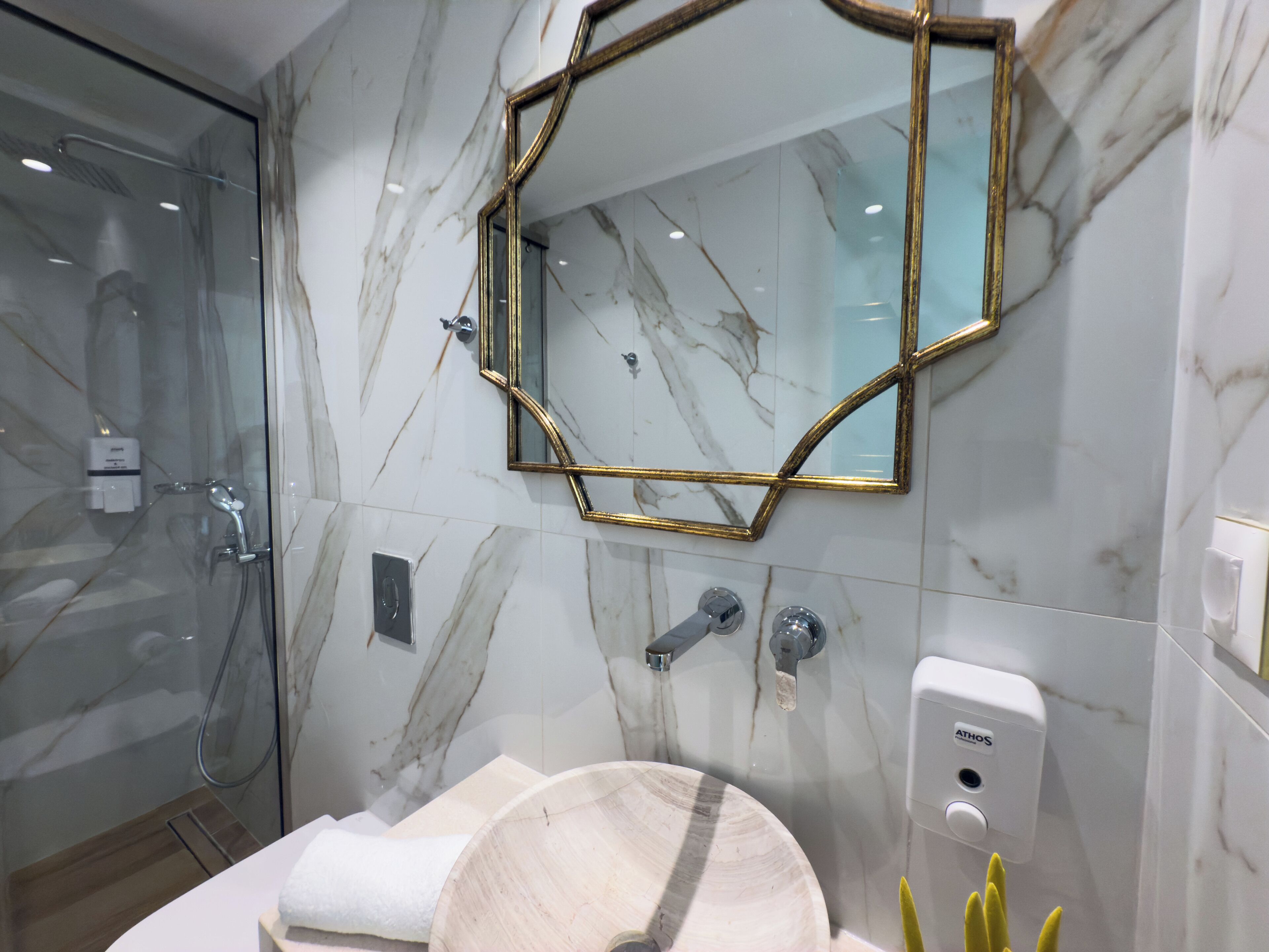 Un baño moderno con pared de mármol, cabina de ducha de vidrio, tocador de madera y espejo con marco dorado.