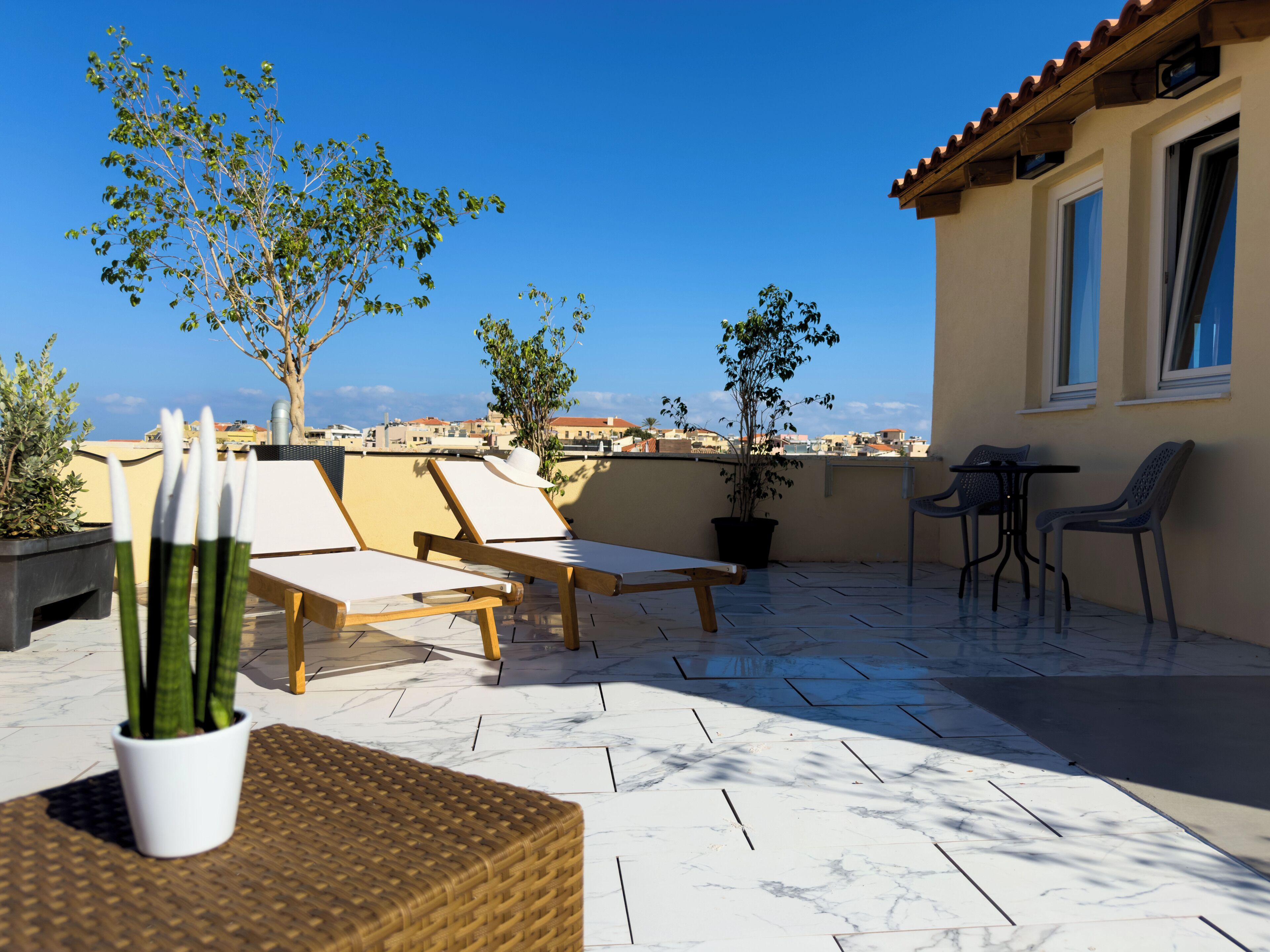 A rooftop terrace with lounge chairs, a small table, potted plants, and a view of the cityscape.