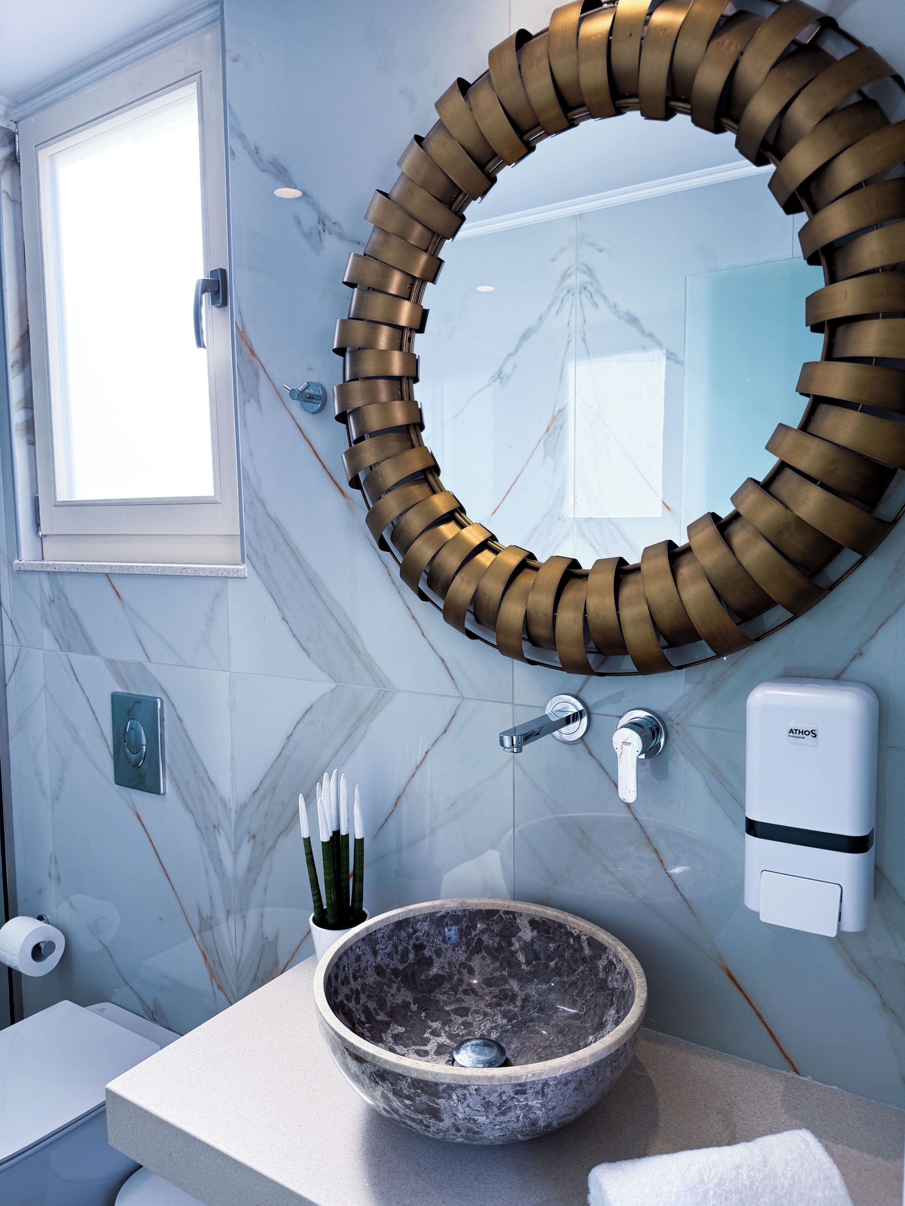 A bathroom with a marble wall, a circular mirror, a sink with a stone basin, a soap dispenser, and a window.