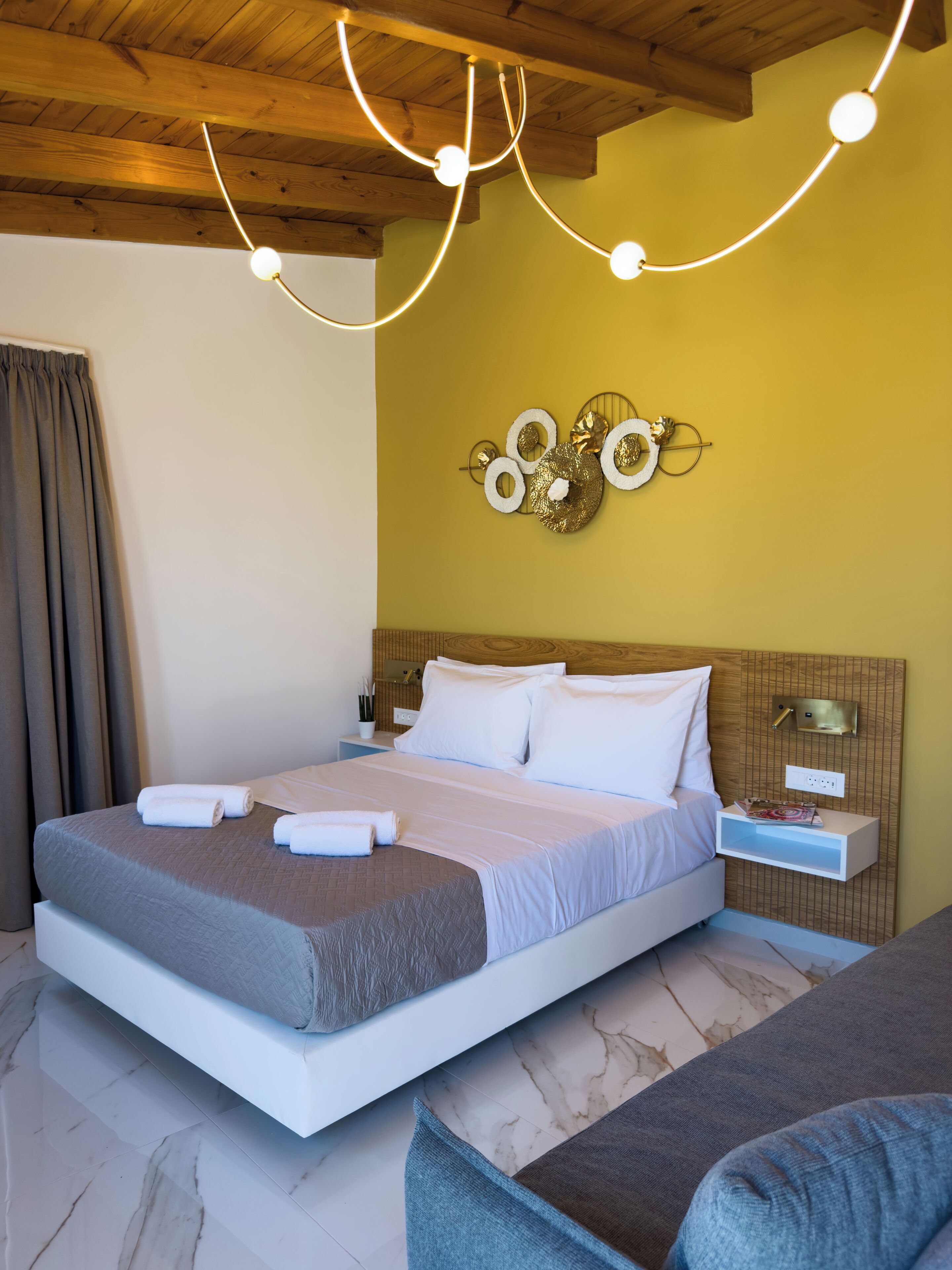 A modern hotel room with a wooden ceiling, a bed with white linens, a nightstand with a lamp, and a decorative wall piece.