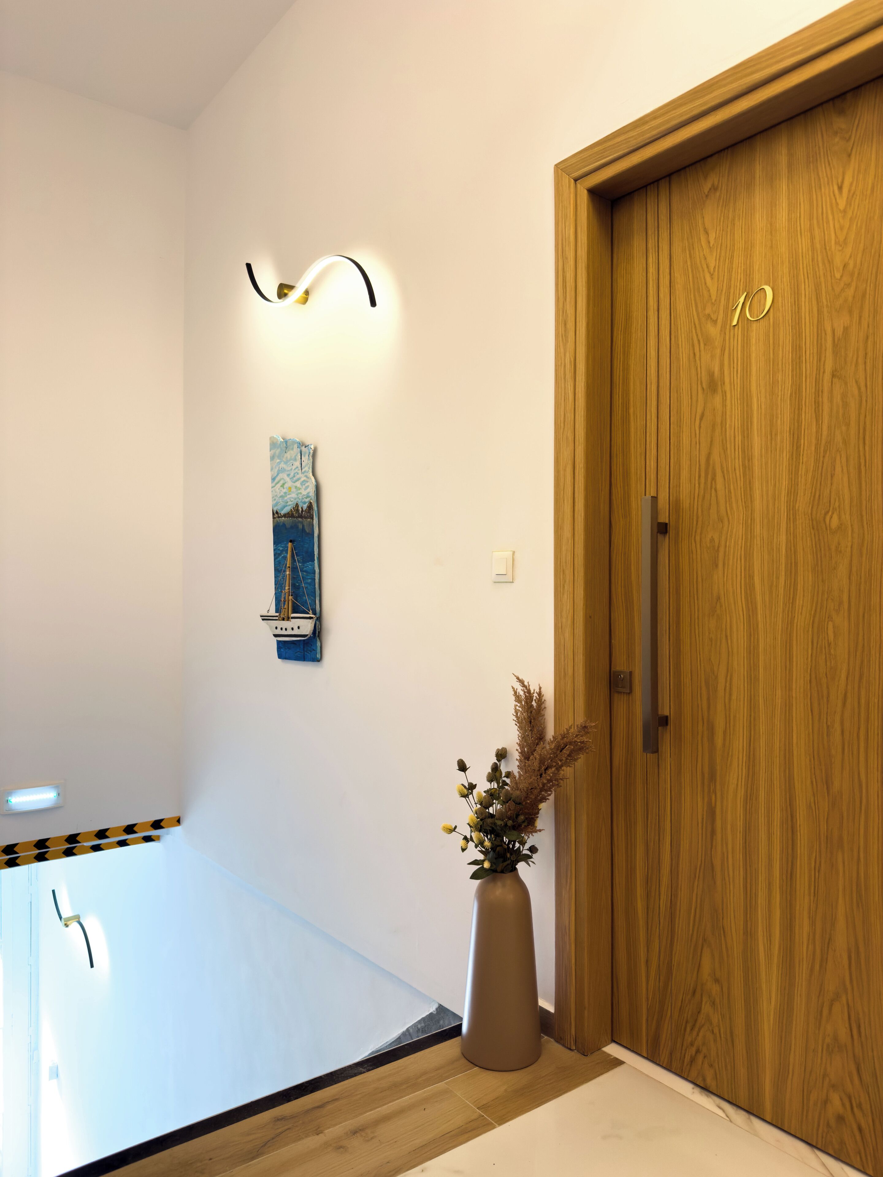 A hallway with a wooden door, a wall-mounted light fixture, a framed artwork, and a vase with dried plants.