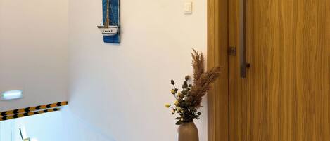 A hallway with a wooden door, a wall-mounted light fixture, a framed artwork, and a vase with dried plants.