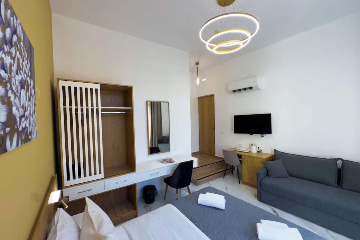 A modern hotel room with a bed, a gray sofa, a desk with a chair, a TV, and a wardrobe.