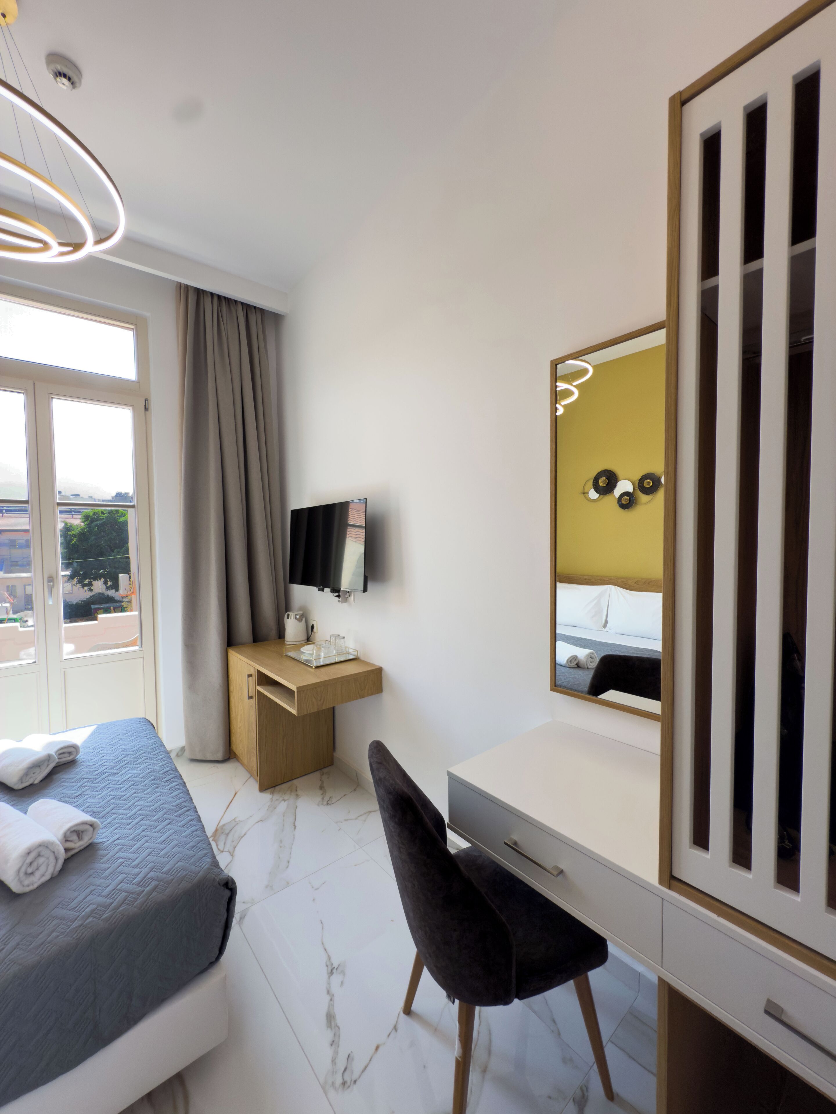 A hotel room with a bed, a desk, a chair, a mirror, and a view of the outside.