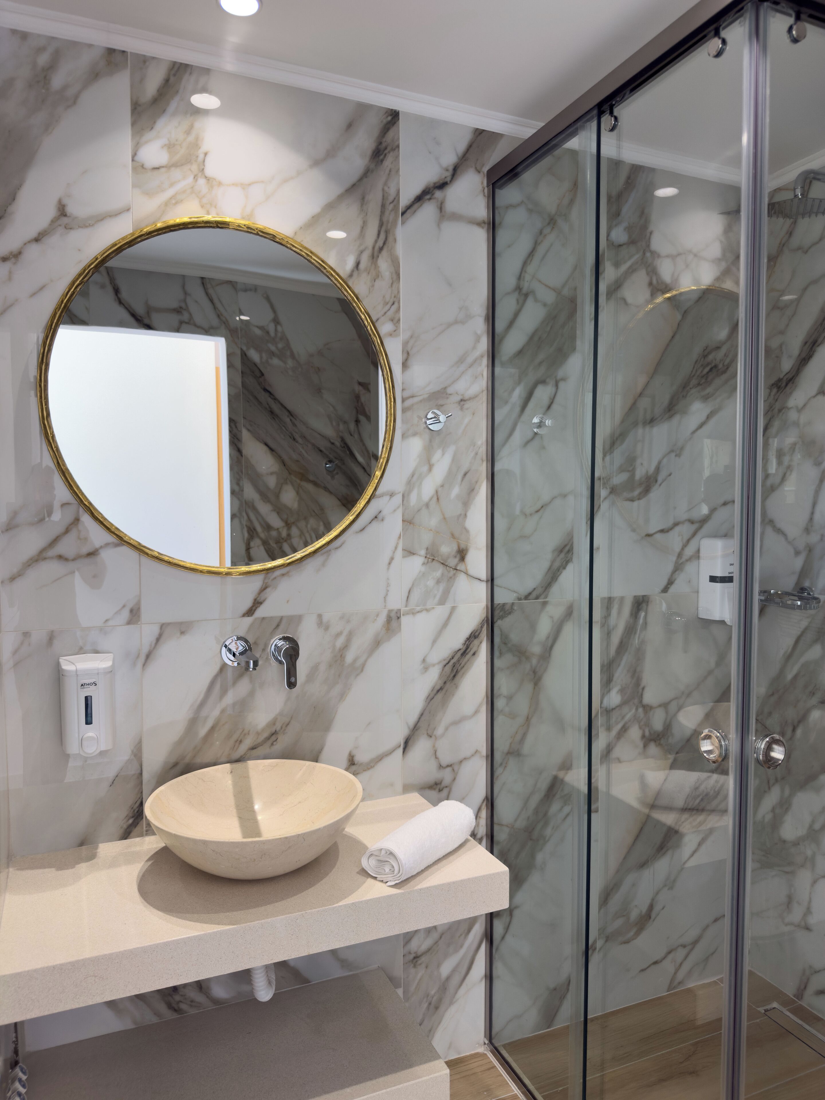 Un baño con pared de mármol, un espejo redondo con marco dorado, un lavabo color beige y una cabina de ducha de vidrio.