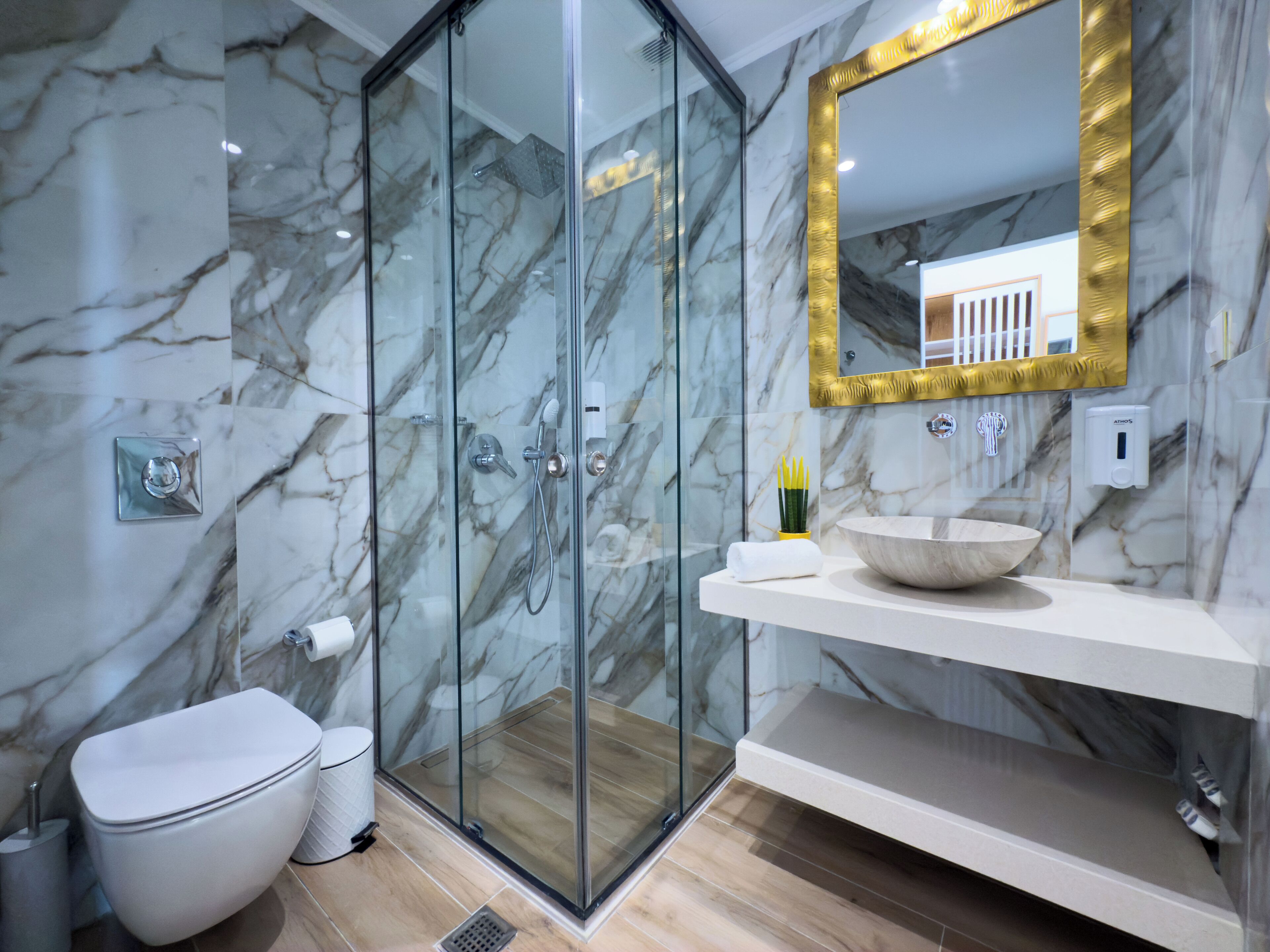 Ein modernes Badezimmer mit einer gläsernen Duschkabine, einer Marmorwand, einem weißen Waschbecken und einem goldfarben gerahmten Spiegel.