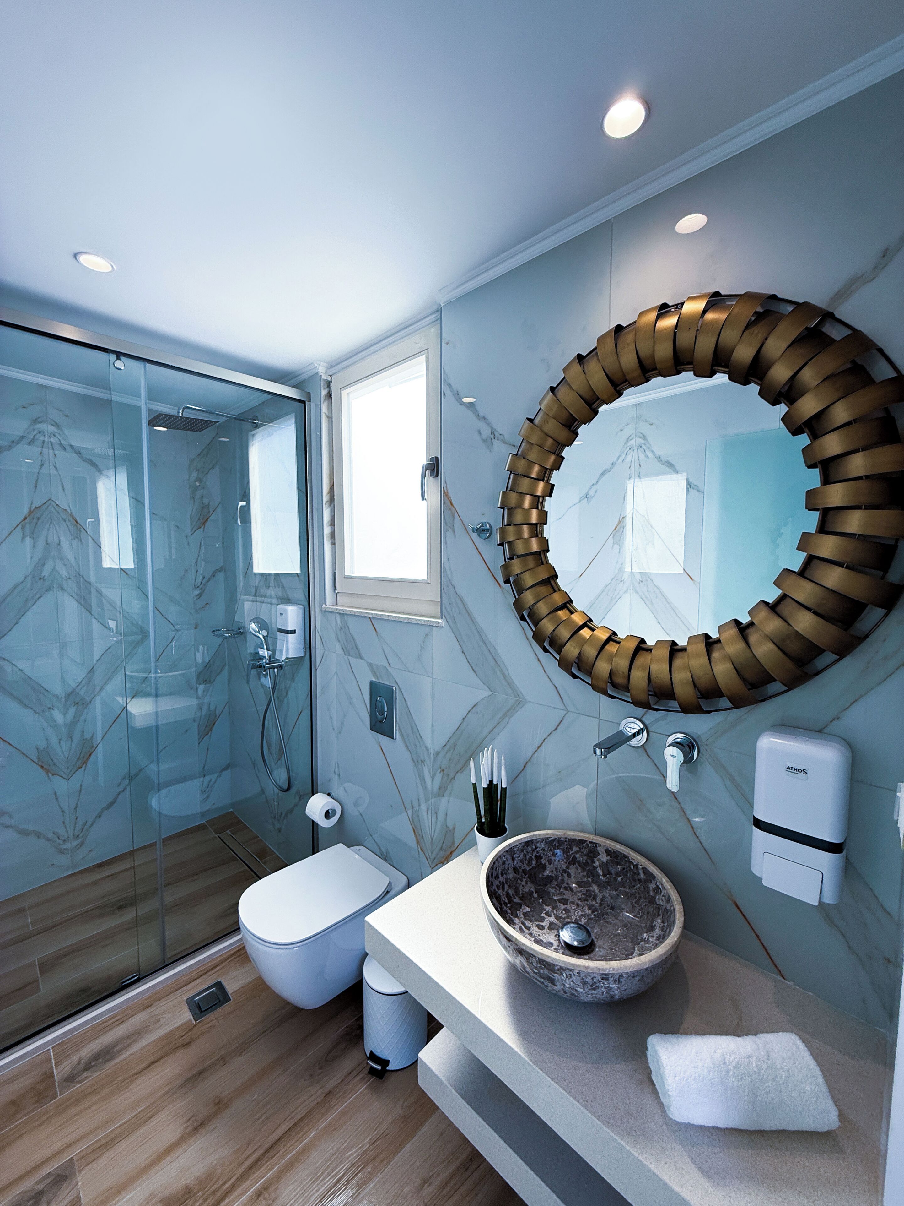 Un baño moderno con cabina de ducha de vidrio, pared con azulejos de mármol, espejo redondo, lavabo con pila de piedra y un inodoro.