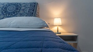 Ein Bett mit blauer Steppdecke und einem gemusterten Kissen, ein Nachttisch mit Lampe sowie eine schlichte Wand.