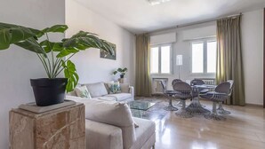 Ein modernes Wohnzimmer mit einer Couch, einem Couchtisch und einem Essbereich mit Stühlen. Eine große Kühlpflanze steht auf einem Marmorsockel.