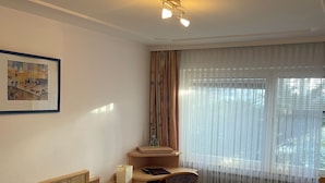 A hotel room with two beds, a desk with a chair, a window with sheer curtains, and a ceiling light. - Gästehaus am Kurpark VS (Villingen-Schwenningen)