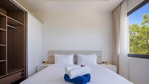 A double bed with white linens and blue towels, a nightstand with a lamp, a wooden wardrobe, and a window with a view of trees. - Choeng mon Modern 2BR Villa (Koh Samui)