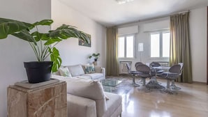 Un salon moderne avec un canapé, une table basse et un coin repas avec des chaises. On y trouve une grande plante en pot, placée sur un piédestal en marbre.