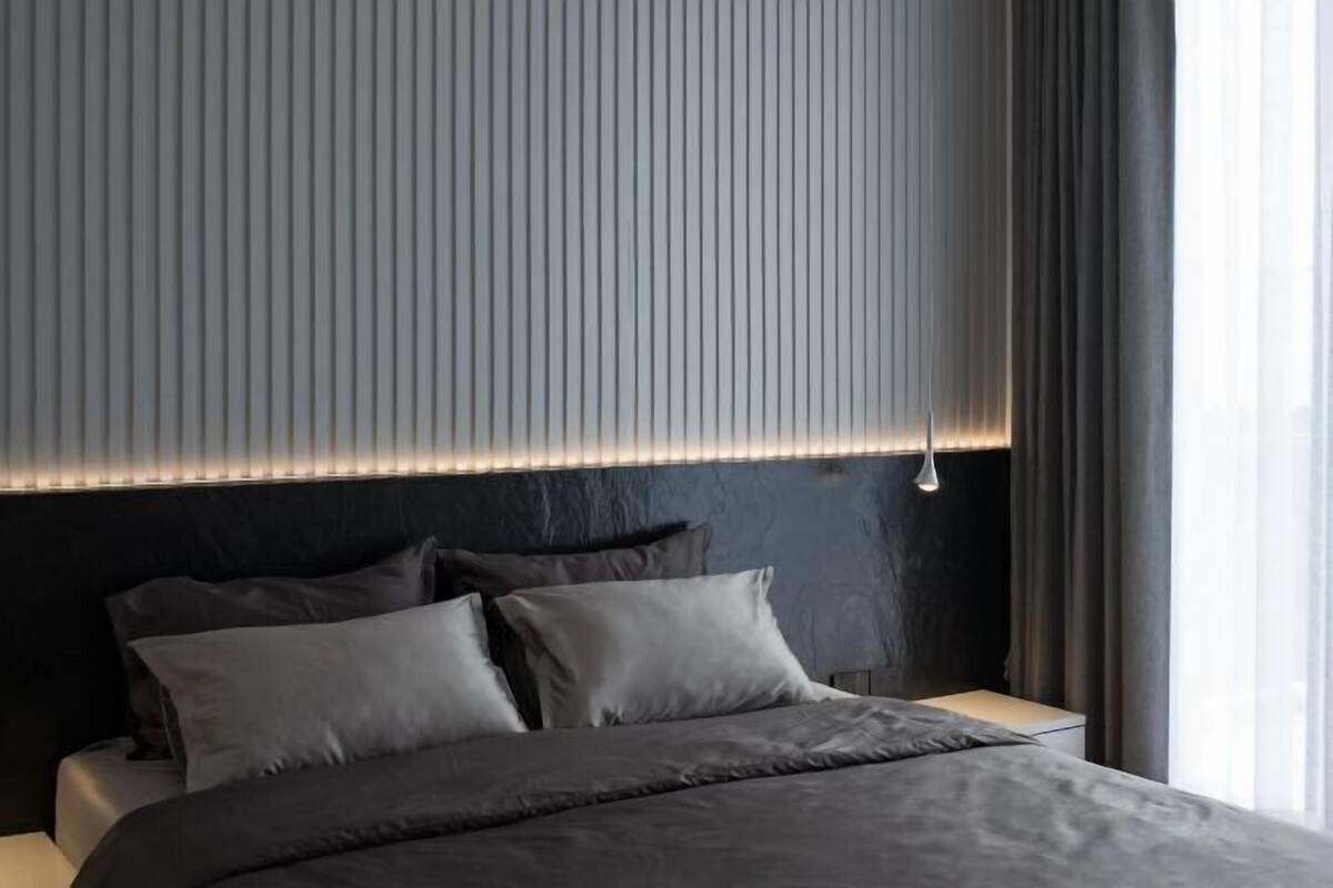 Een moderne slaapkamer met een donker hoofdbord, een bed met grijze beddengoed en een raam met gordijnen.