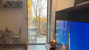 A modern living room with a large flat-screen TV mounted on a wooden console, a dining area with a table and chairs, and a view of a cityscape through an open door.
