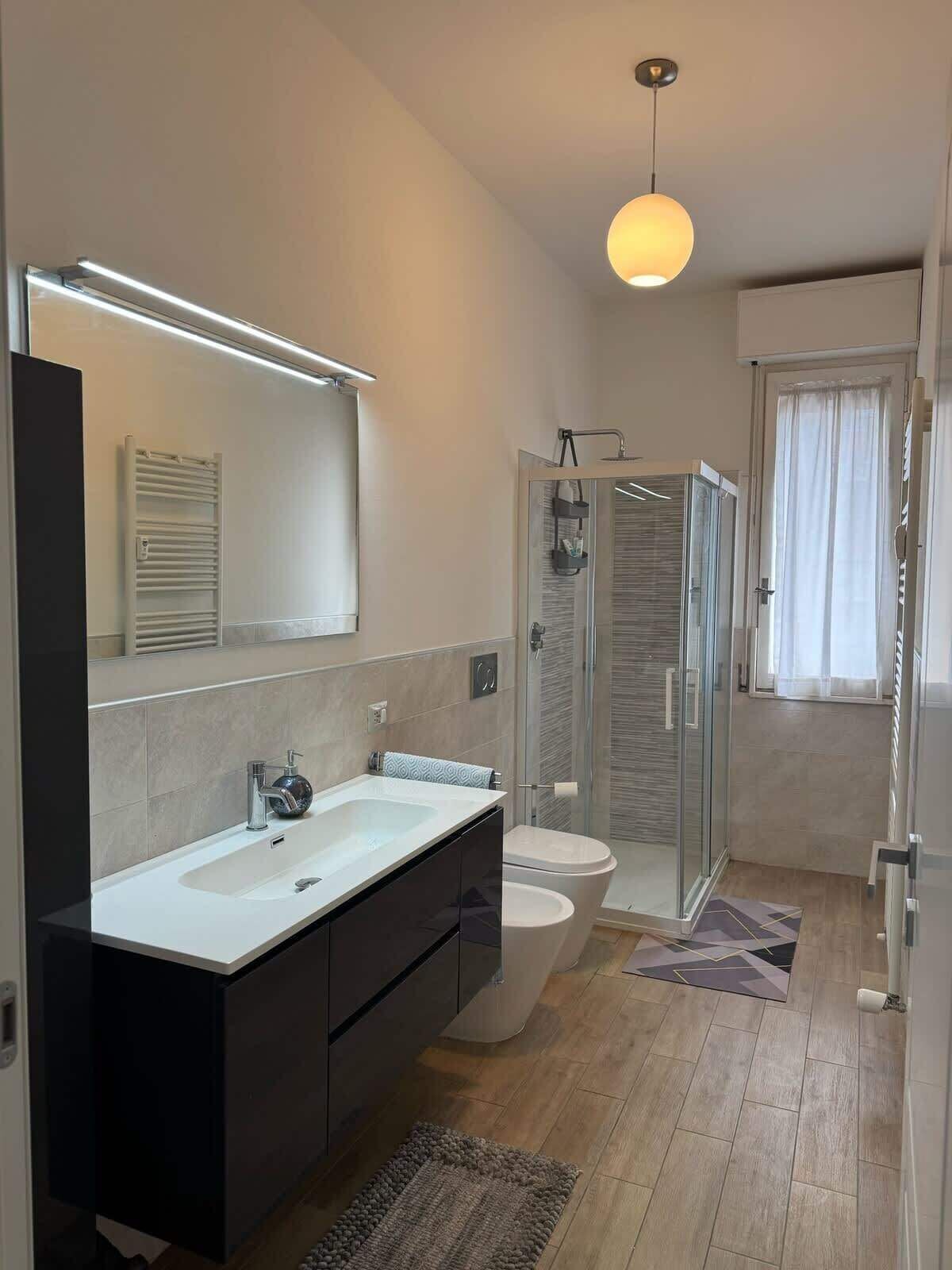A modern bathroom with a large mirror, a white sink, a black cabinet, a glass shower enclosure, and a toilet.