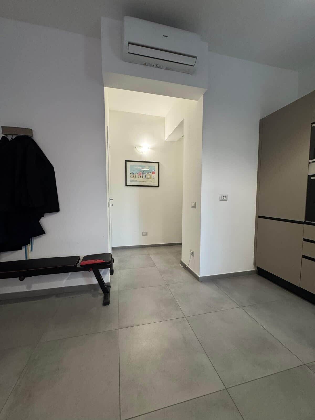 A room with a bench, a wall-mounted air conditioner, a framed picture, and a kitchenette.