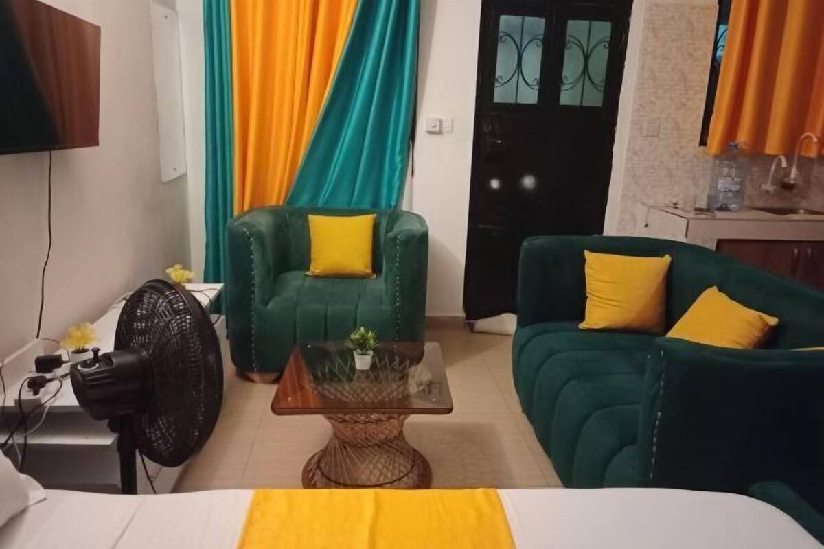 A living room with a green sofa, a green armchair, a glass coffee table, and a black fan.