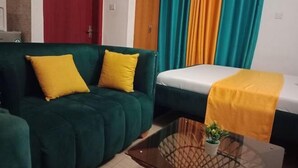 Ein Zimmer mit einem grünen Sofa, gelben Kissen, einem weißen Bett und einem Glastischchen mit einer Pflanze.