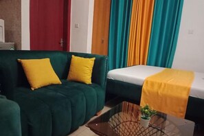 A room with a green sofa, a bed with a yellow blanket, and a small table with a plant. - Royal Studio Apartments (Bamburi)