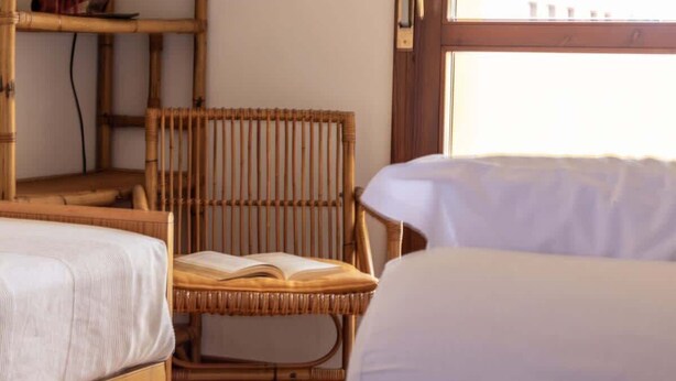 A room with two wooden beds, a wicker chair with an open book, and a window with a view.