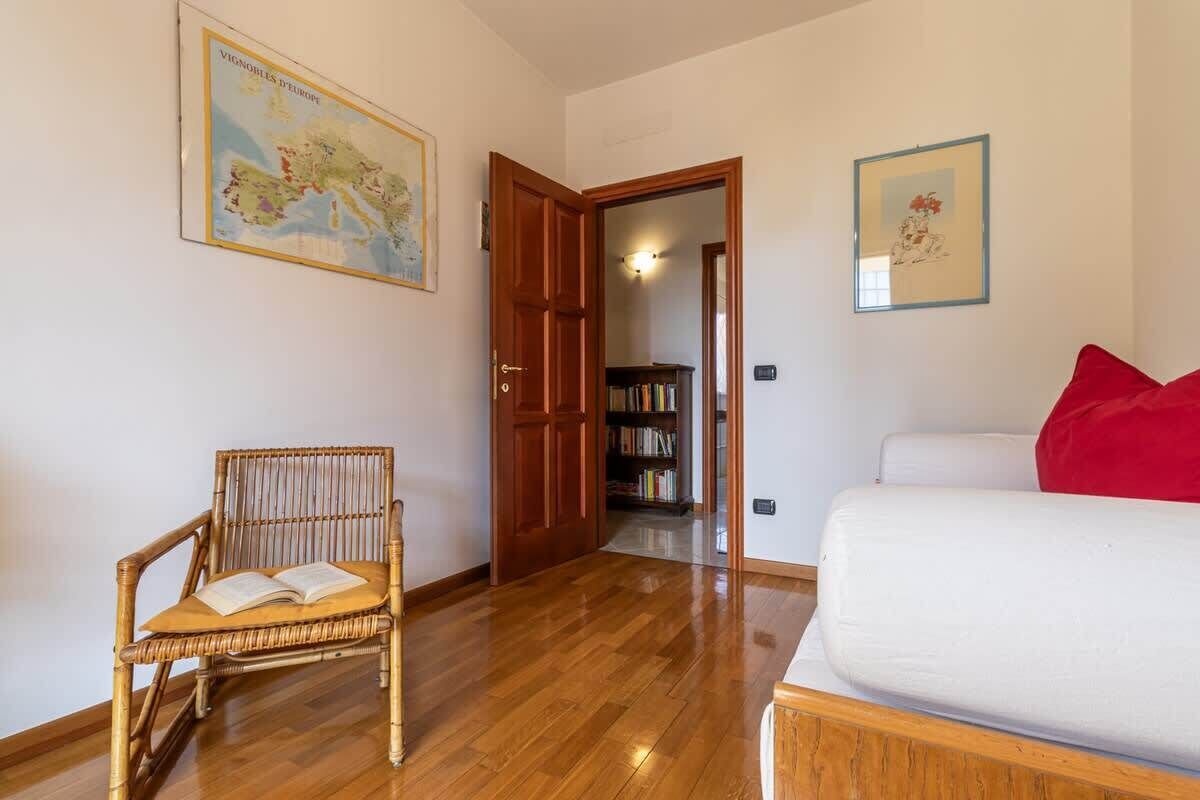Una habitació amb un sòl de fusta, un sofà blanc amb un coixí vermell, una cadira de vimetlla amb un llibre, una porta de fusta i una biblioteca al fons.
