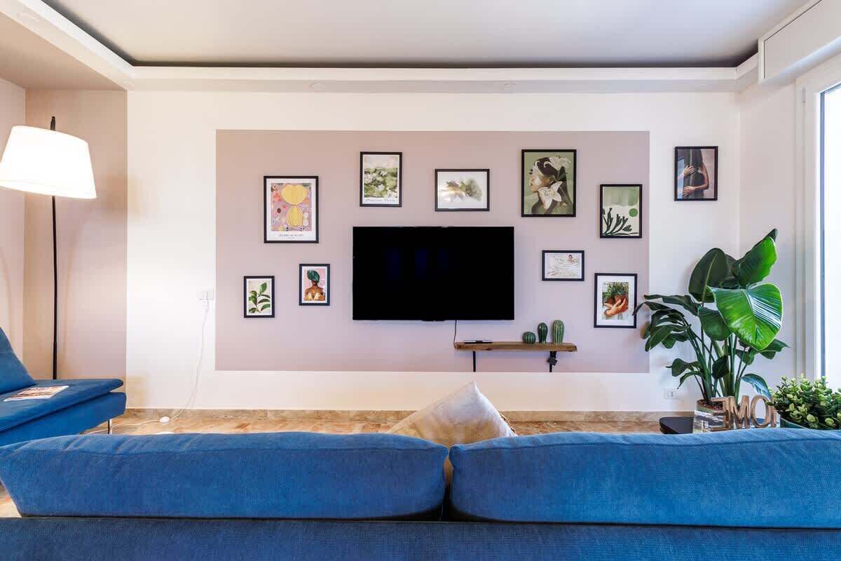 A living room with a large flat-screen TV mounted on the wall, surrounded by framed artwork. There is a blue sofa with cushions, a floor lamp, and a large green plant.