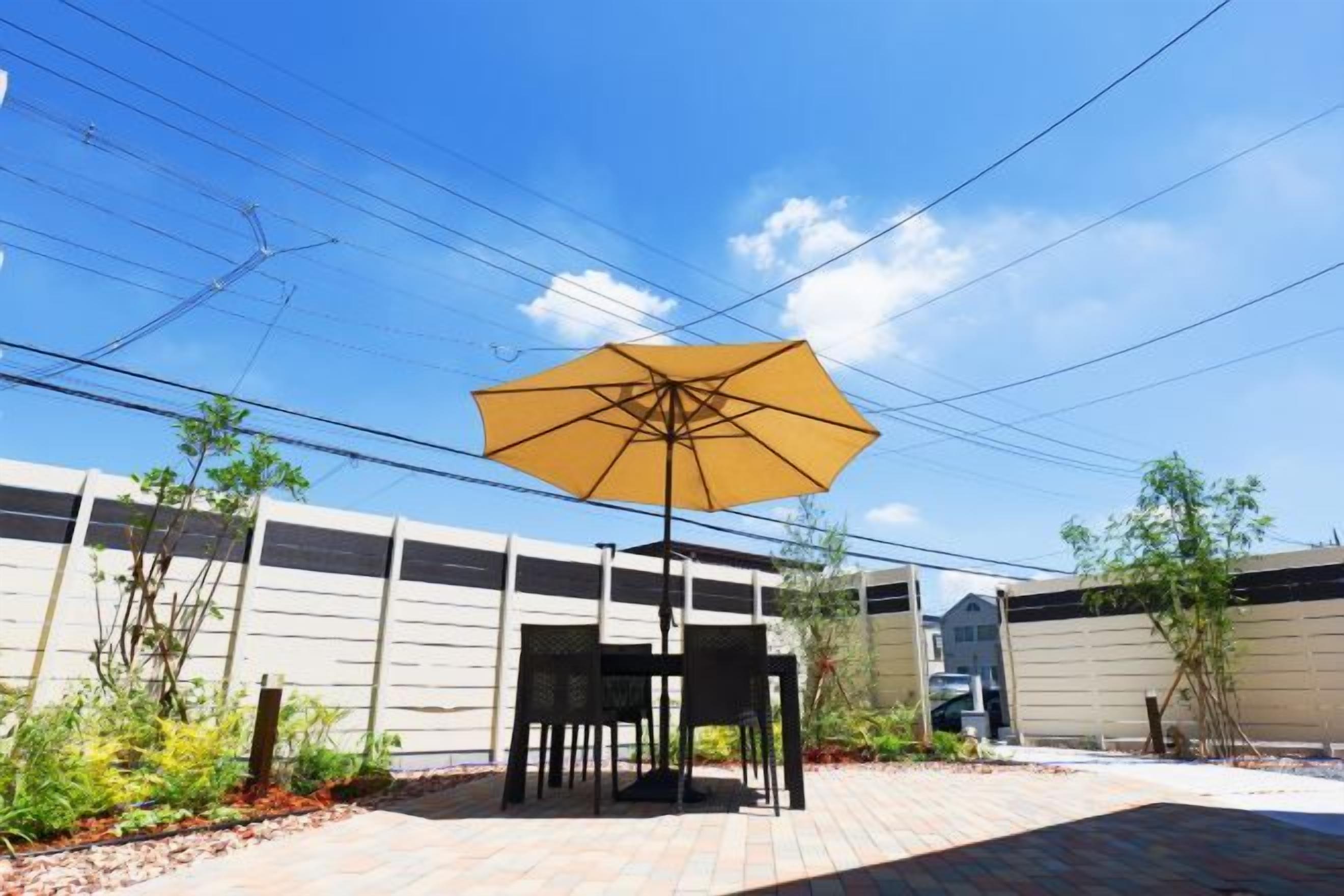An outdoor patio area with a table and chairs under a large umbrella, surrounded by plants and a brick floor.
