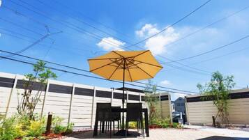 An outdoor patio area with a table and chairs under a large umbrella, surrounded by plants and a brick floor.