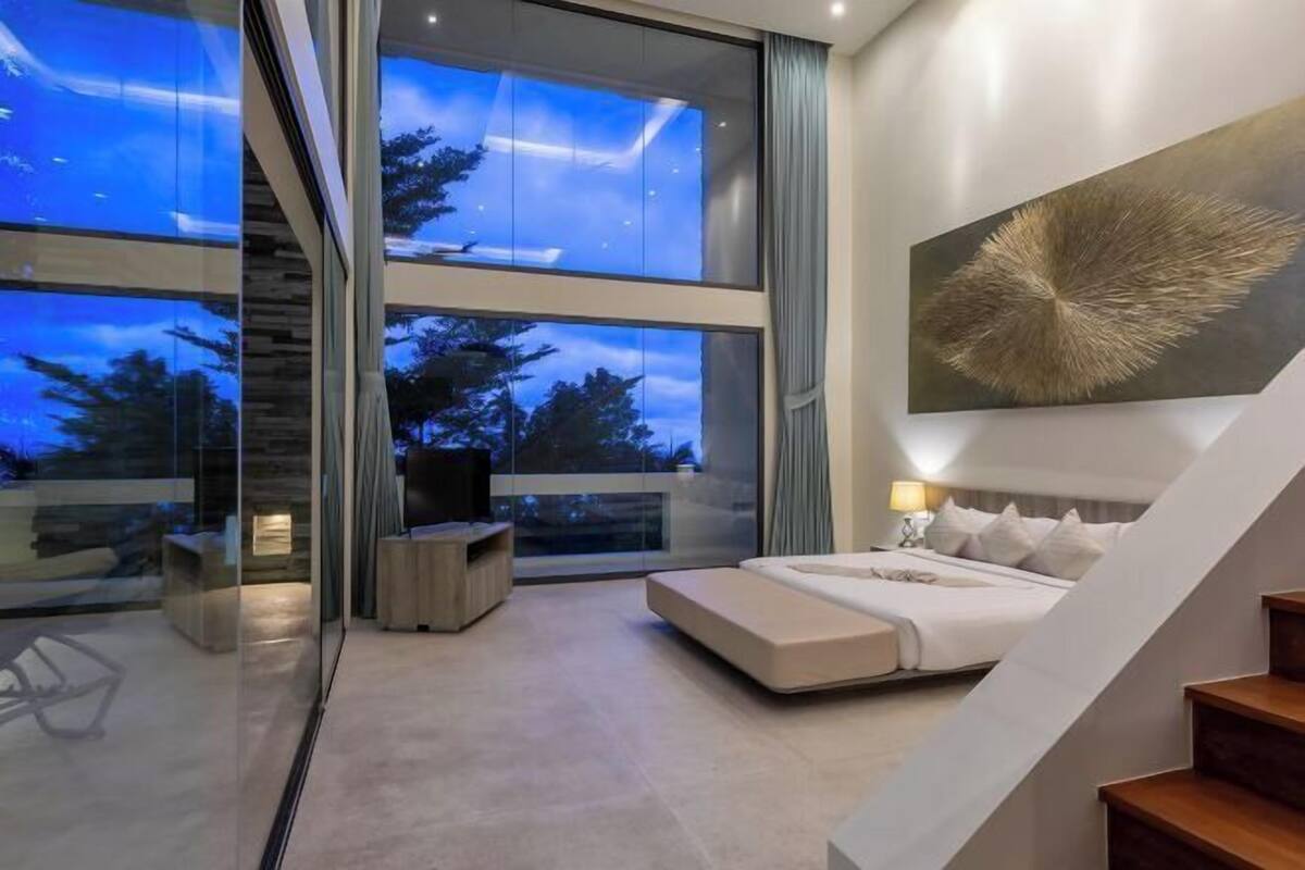 A modern bedroom with a large bed, a view through floor-to-ceiling windows, and a staircase.