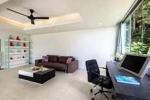 A modern living room with a sofa, coffee table, and a desk with a laptop. There is a bookshelf with colorful figurines and a ceiling fan. - Villa Spice Samui (Koh Samui)