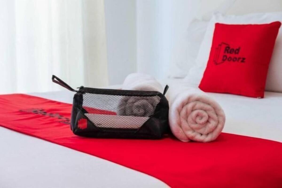 Una cama bien hecha con una manta roja y una toalla enrollada. Una bolsa de malla negra está colocada sobre la cama.